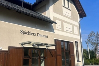 Budynek o jasnej elewacji z ciemnym dachem, dużymi drewnianymi drzwiami i przeszklonymi oknami na parterze. Przed budynkiem znajduje się kamienny podjazd, a w tle widać drzewa i niebo.